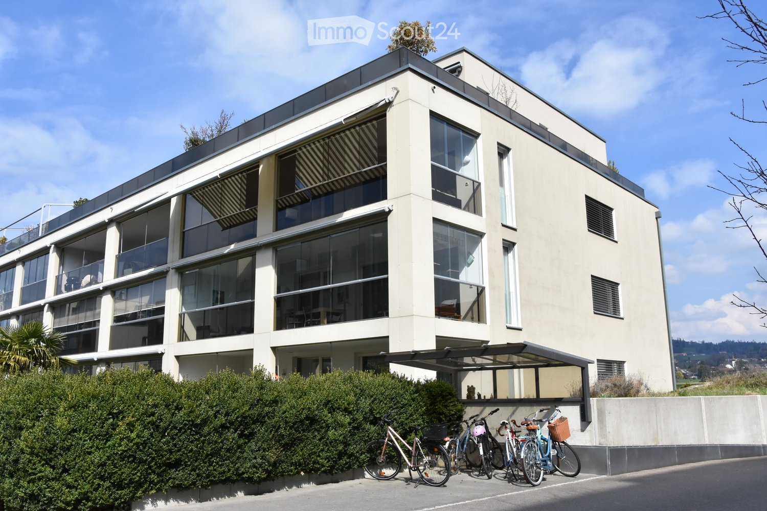 Modern multi-story residential building with large windows, balconies, and a flat roof. Bicycles are parked in front of the building.