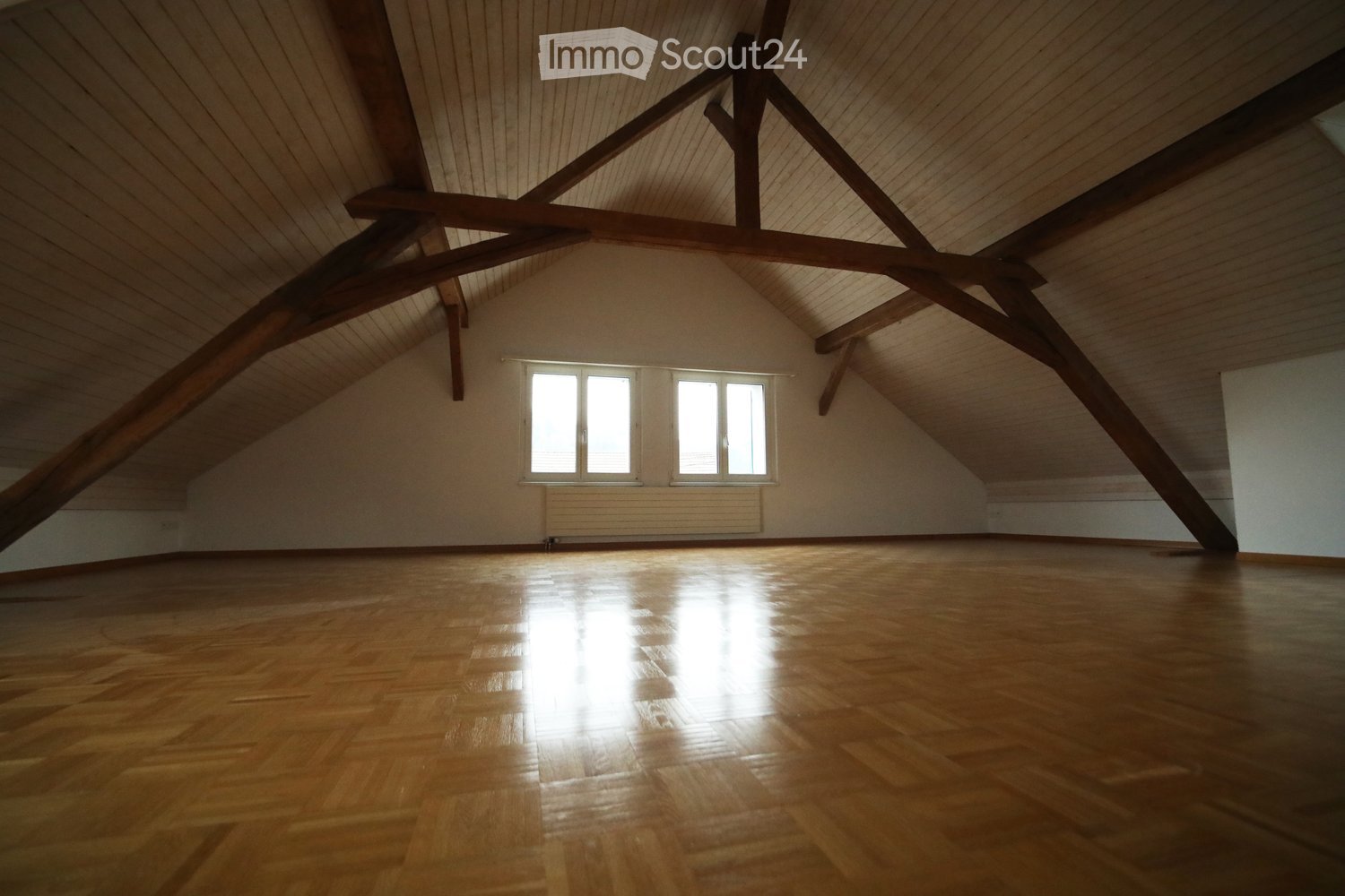 Empty attic room with wooden floor, exposed wooden beams, and large windows allowing natural light to enter.