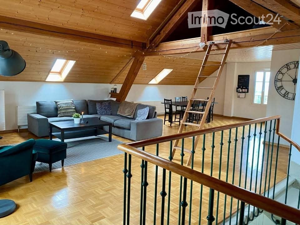 Open-plan living and dining area with wooden beams, skylights, grey sofa, coffee table, dining table, and a staircase leading to a loft area.