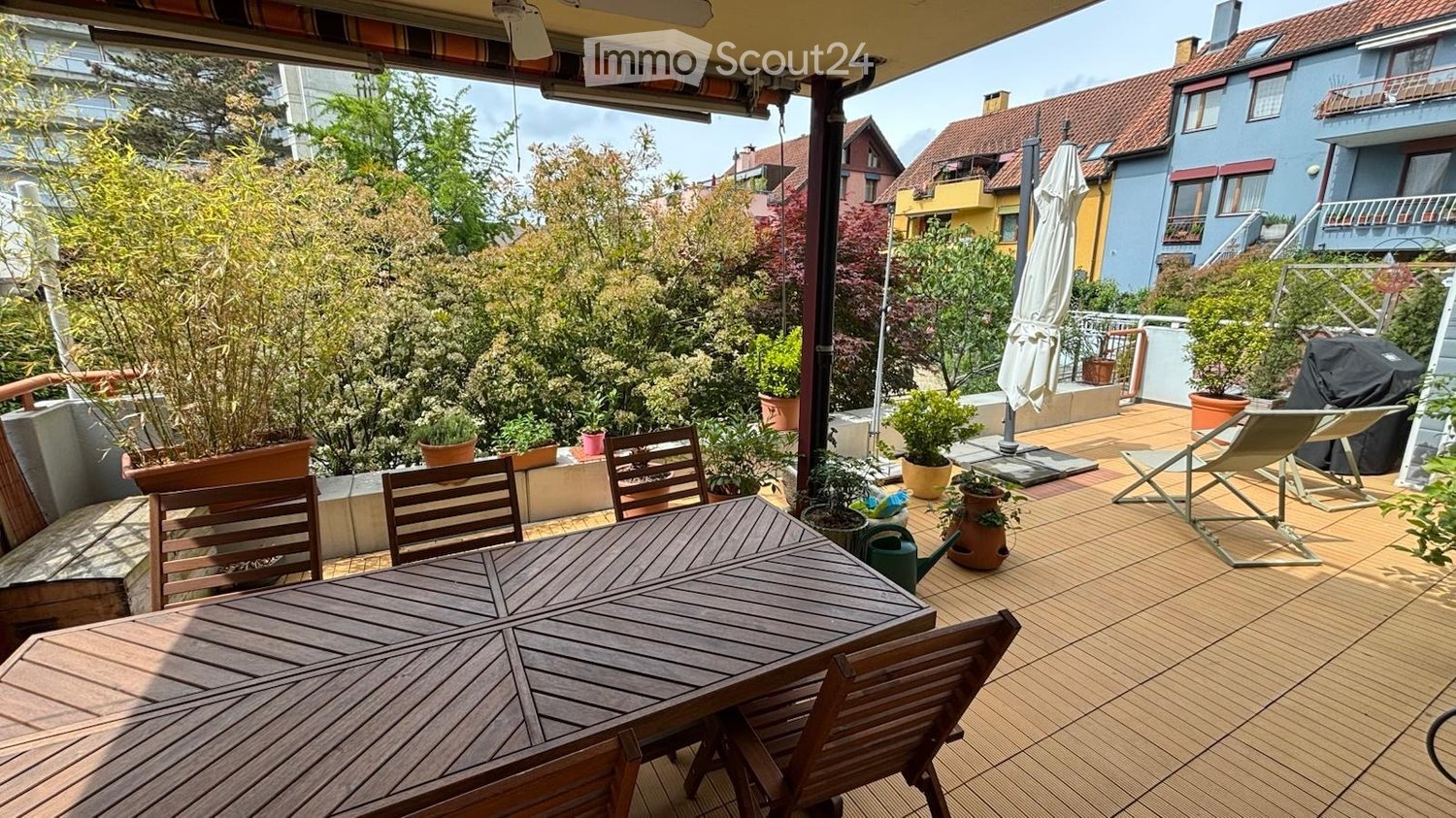 Balcony / Terrace. Wooden deck with a table, chairs, umbrella, and potted plants, overlooking a garden and neighboring houses.