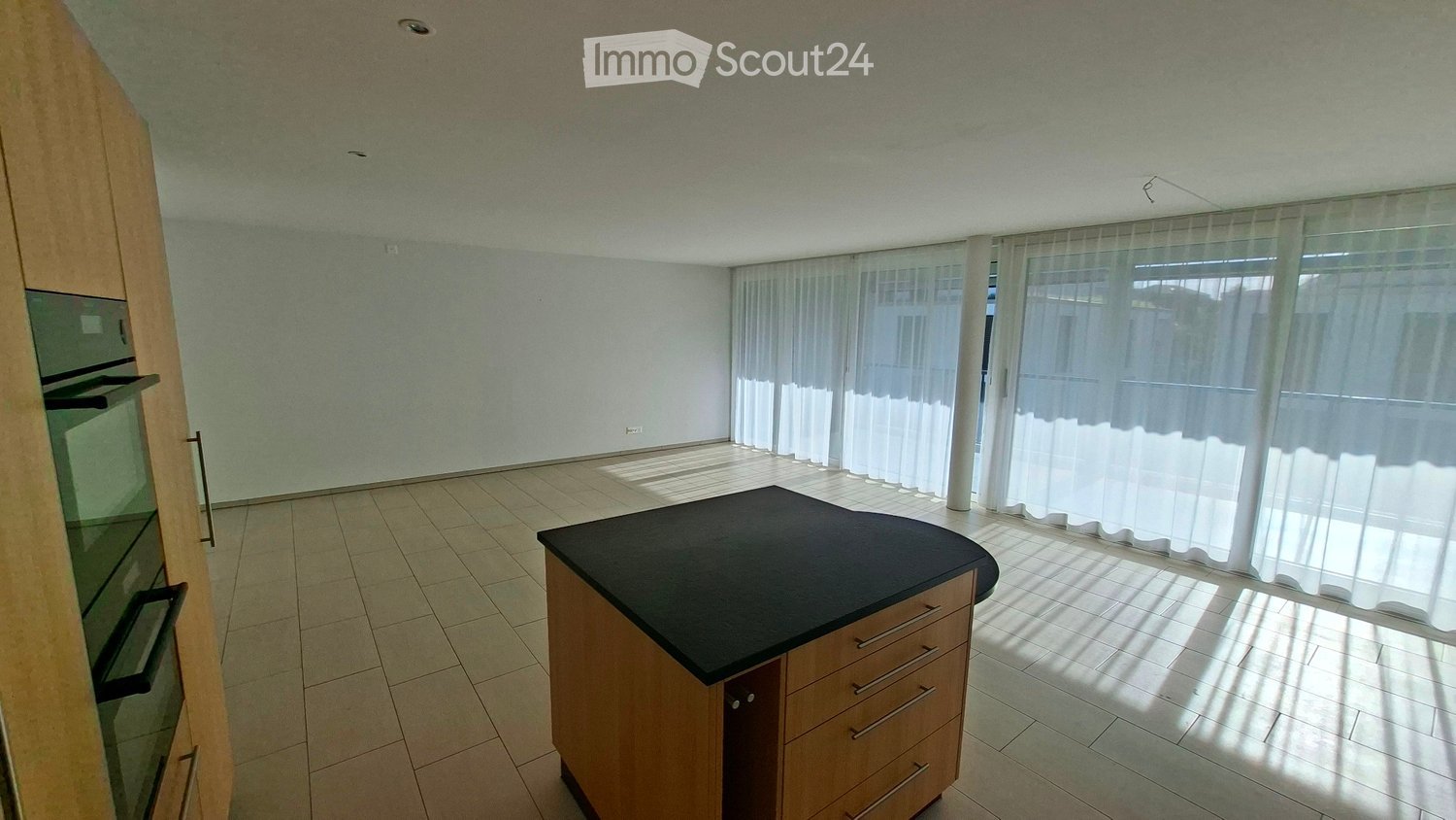 Bright, unfurnished kitchen with a central island and built-in oven, large windows with white curtains, tiled floor.