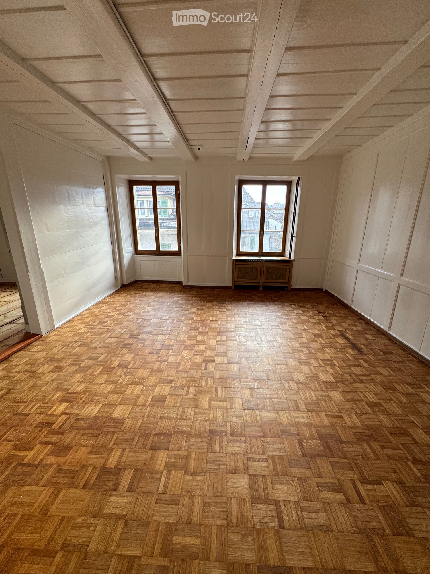 Empty room with wooden herringbone flooring, white walls, and two windows with brown frames. The ceiling has exposed wooden beams.
