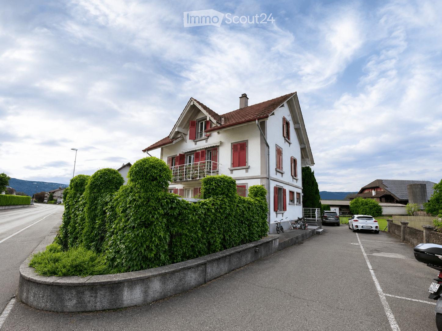 Two-story house with red shutters, white walls, and a brown roof, situated on a quiet street with a well-maintained hedge and a driveway with parked cars.