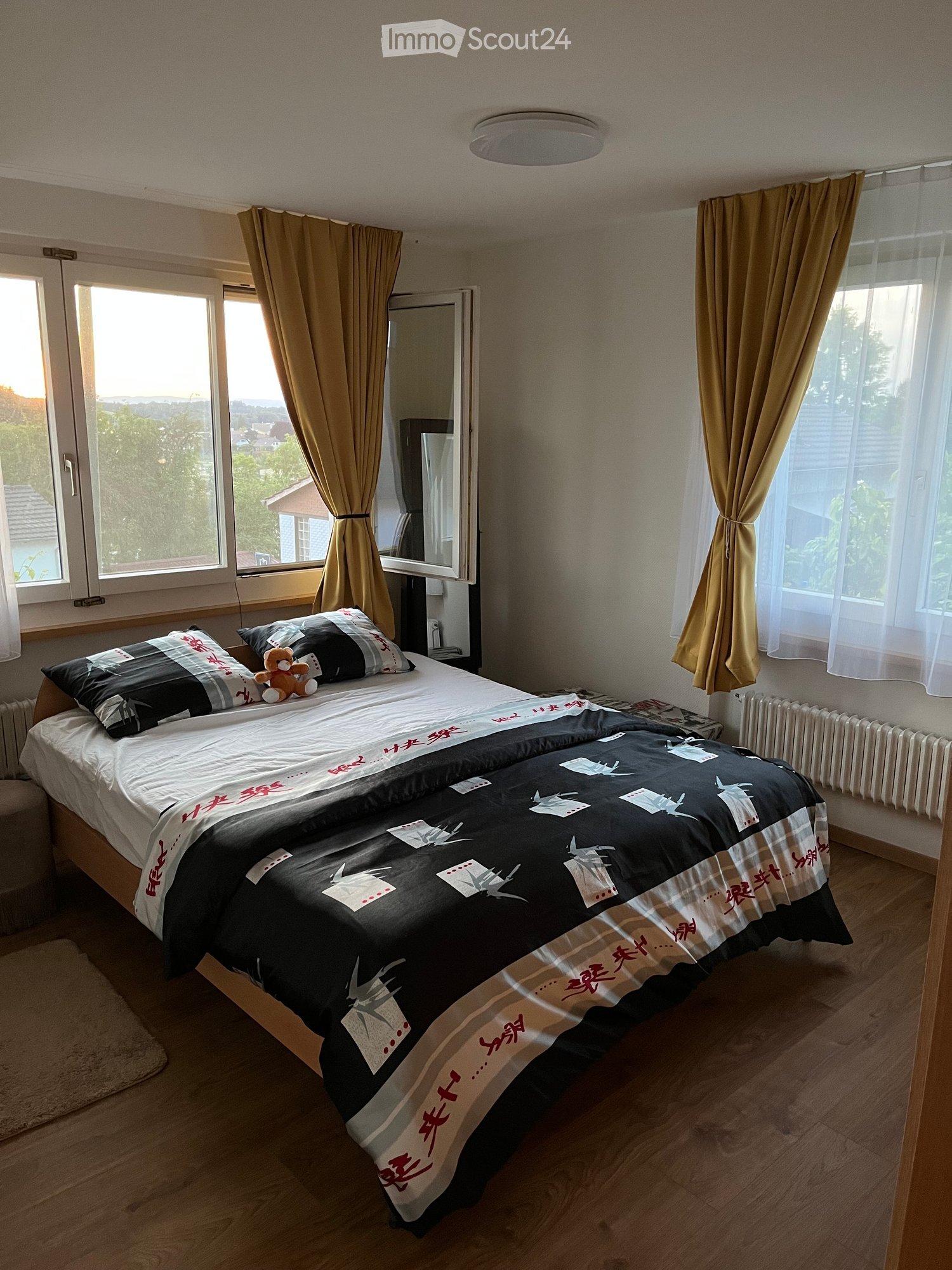 Well-lit bedroom with wooden flooring, large windows with curtains, a double bed with decorative bedding, a teddy bear, and a mirror on a stand.