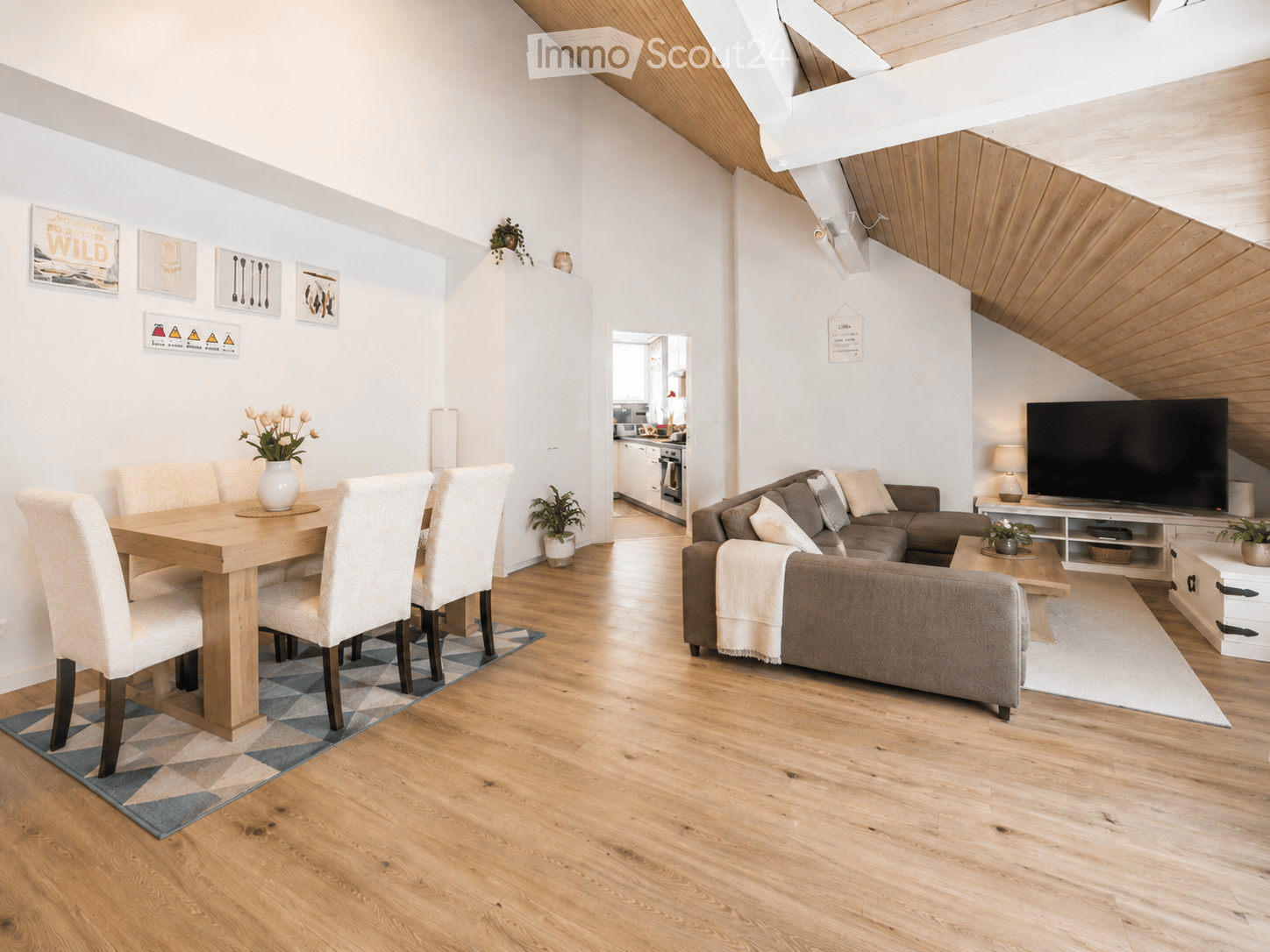 Open-plan living and dining area with wooden flooring, a grey sectional sofa, a wooden dining table with four chairs, a television on a stand, and decorative vases with flowers.