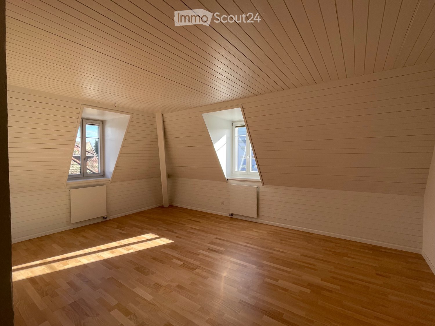 Empty attic room with wooden walls, ceiling, and floor, two windows, and radiators.