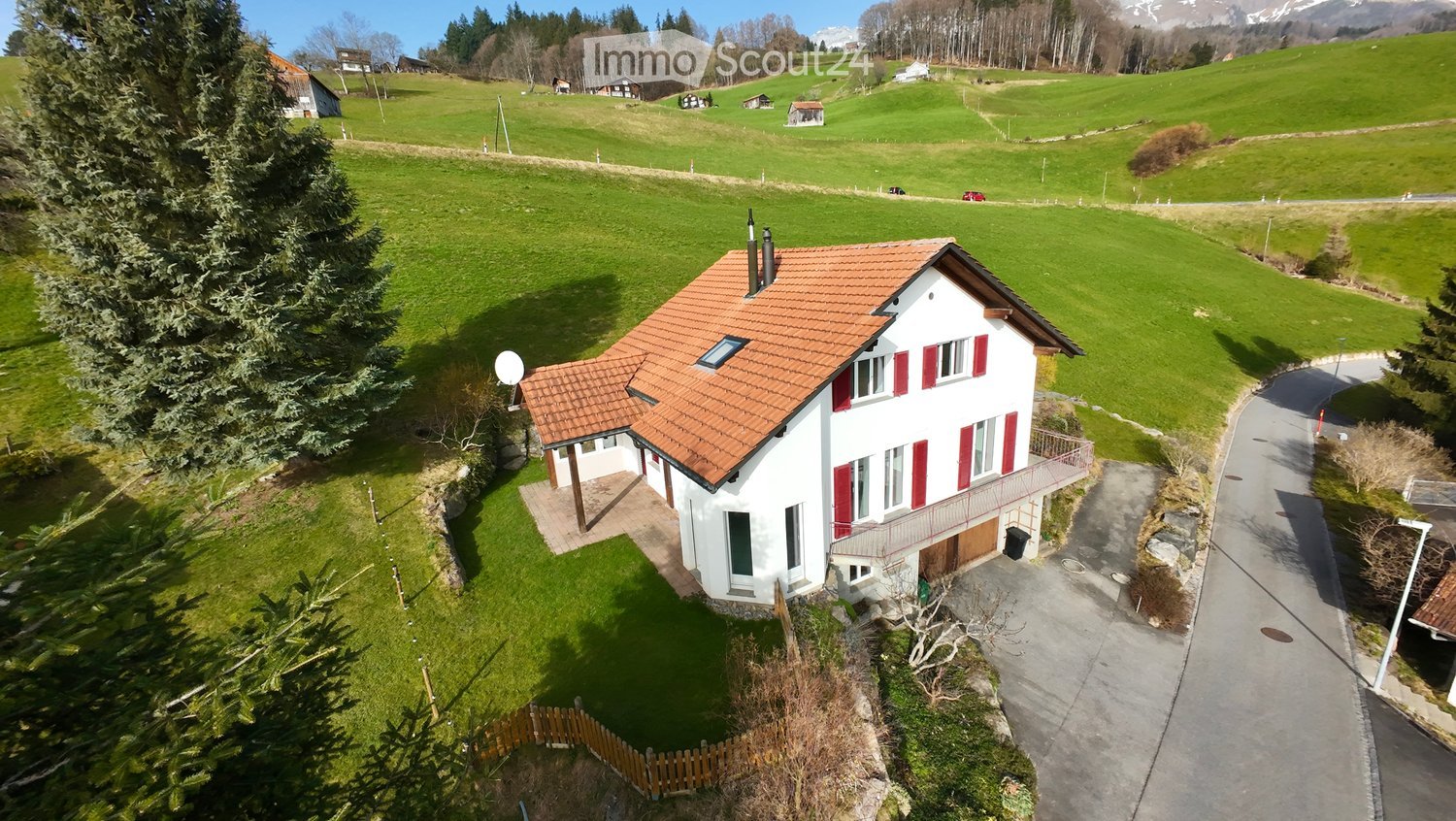 A two-story white house with red shutters and a red-tiled roof, situated in a green, hilly landscape with scattered trees and other houses in the background.