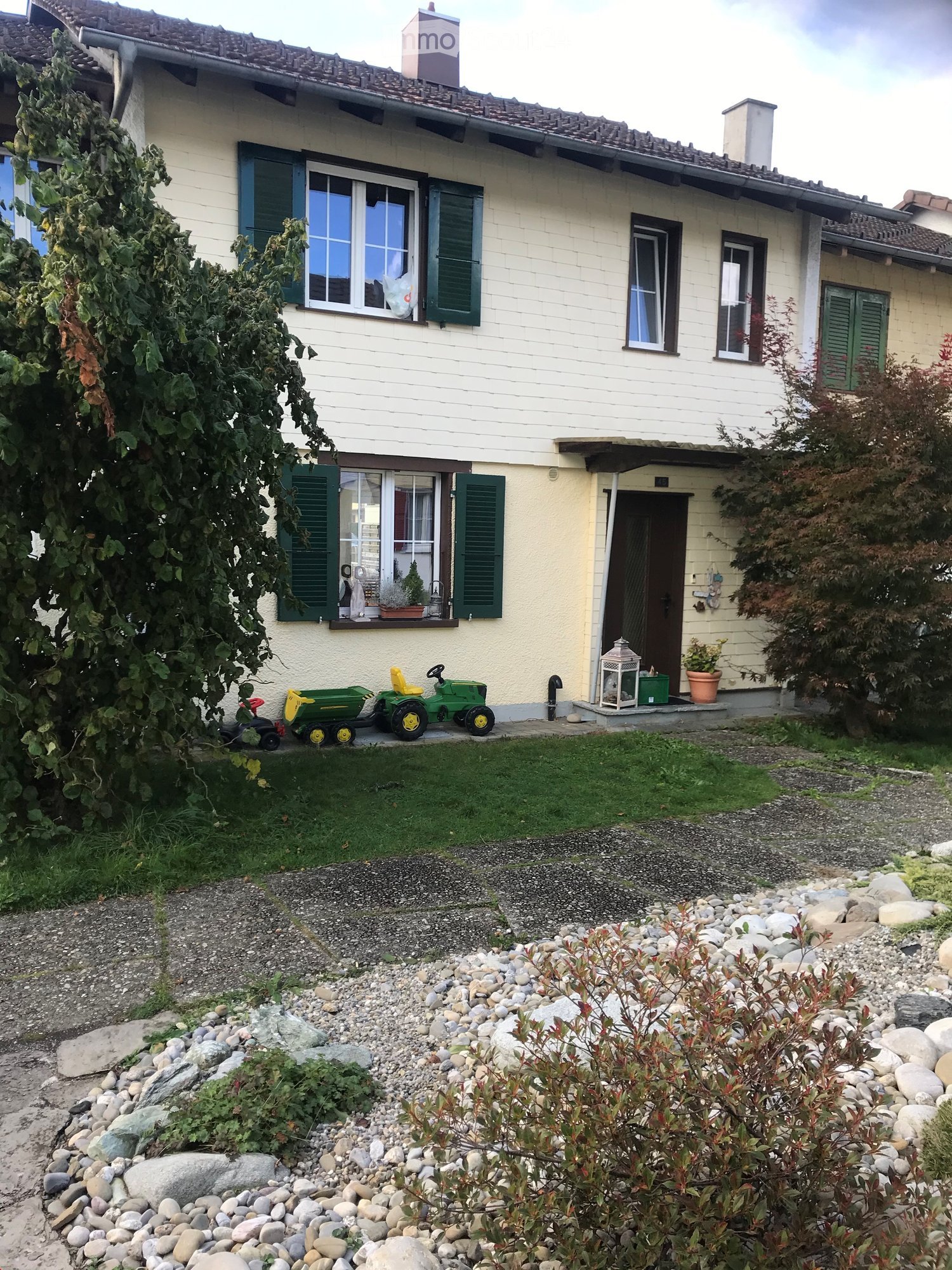 House. Two-story house with light yellow siding, green shutters, and multiple windows. The front yard features a small lawn, a gravel pathway, and decorative rocks. A green toy tractor and a small play vehicle are parked on the grass near the house.