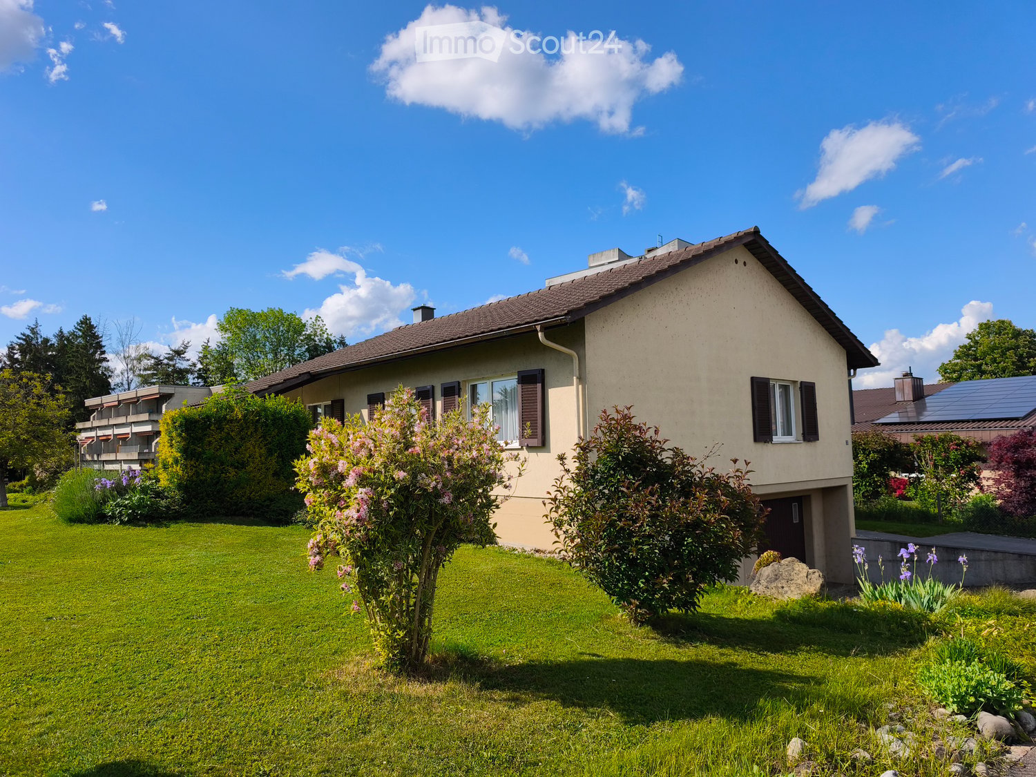 Single-story house with beige exterior, brown roof, multiple windows with shutters, surrounded by a well-maintained lawn and garden with flowering shrubs and trees.