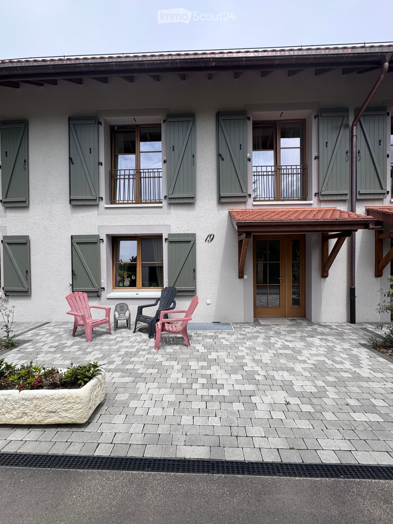 Paved outdoor area with four chairs, a small table, and a planter near a two-story house with shutters and a covered entrance.