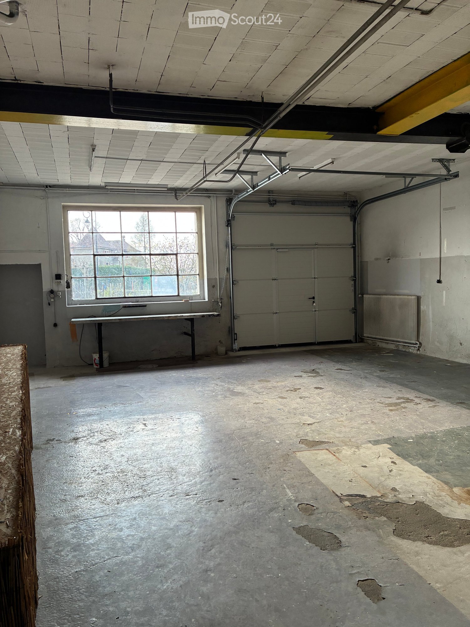 Empty room with a large window, a table, a white bucket, a radiator, and a garage door.