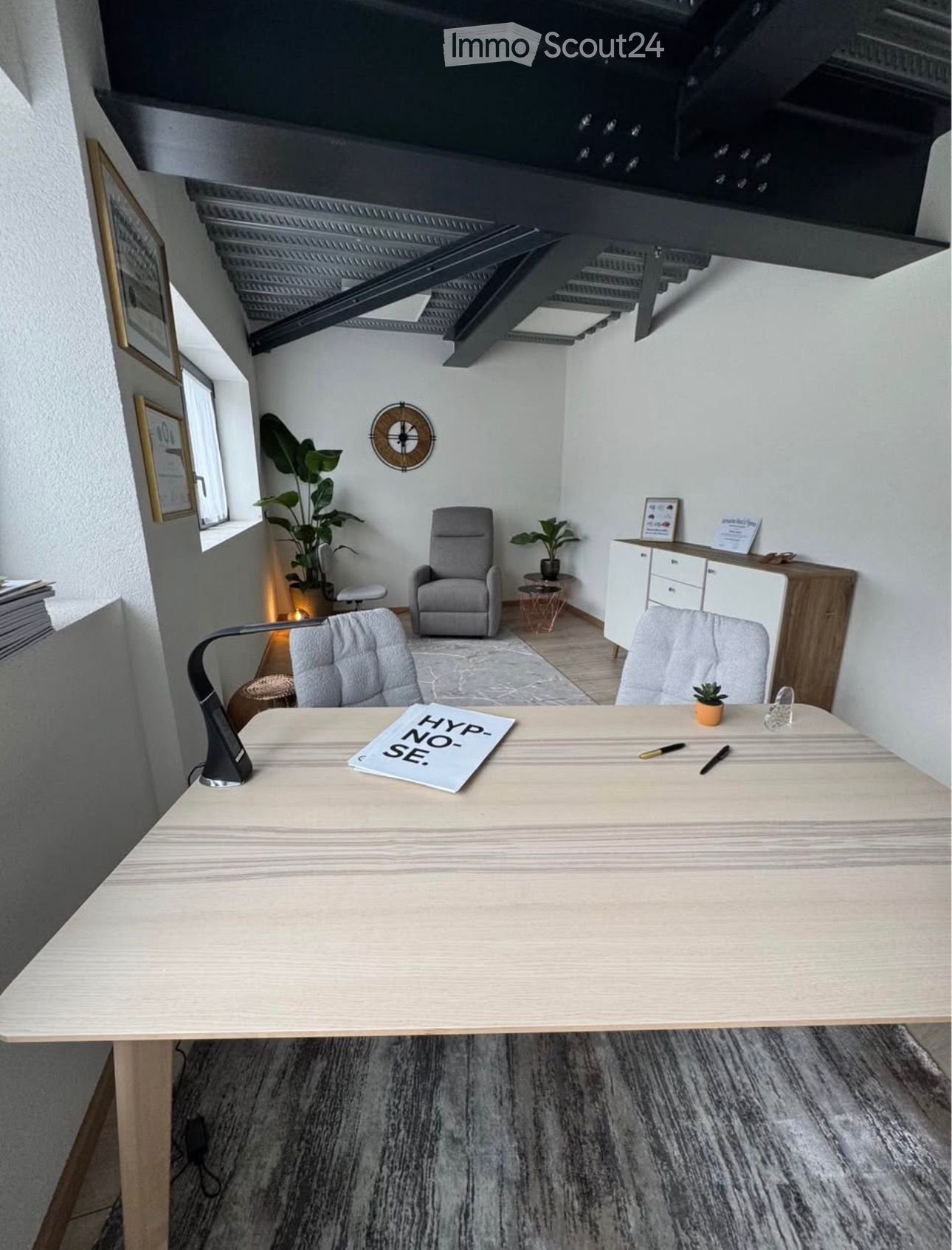 Modern office space with a large desk, ergonomic chairs, a lamp, potted plants, and a cabinet with framed certificates.