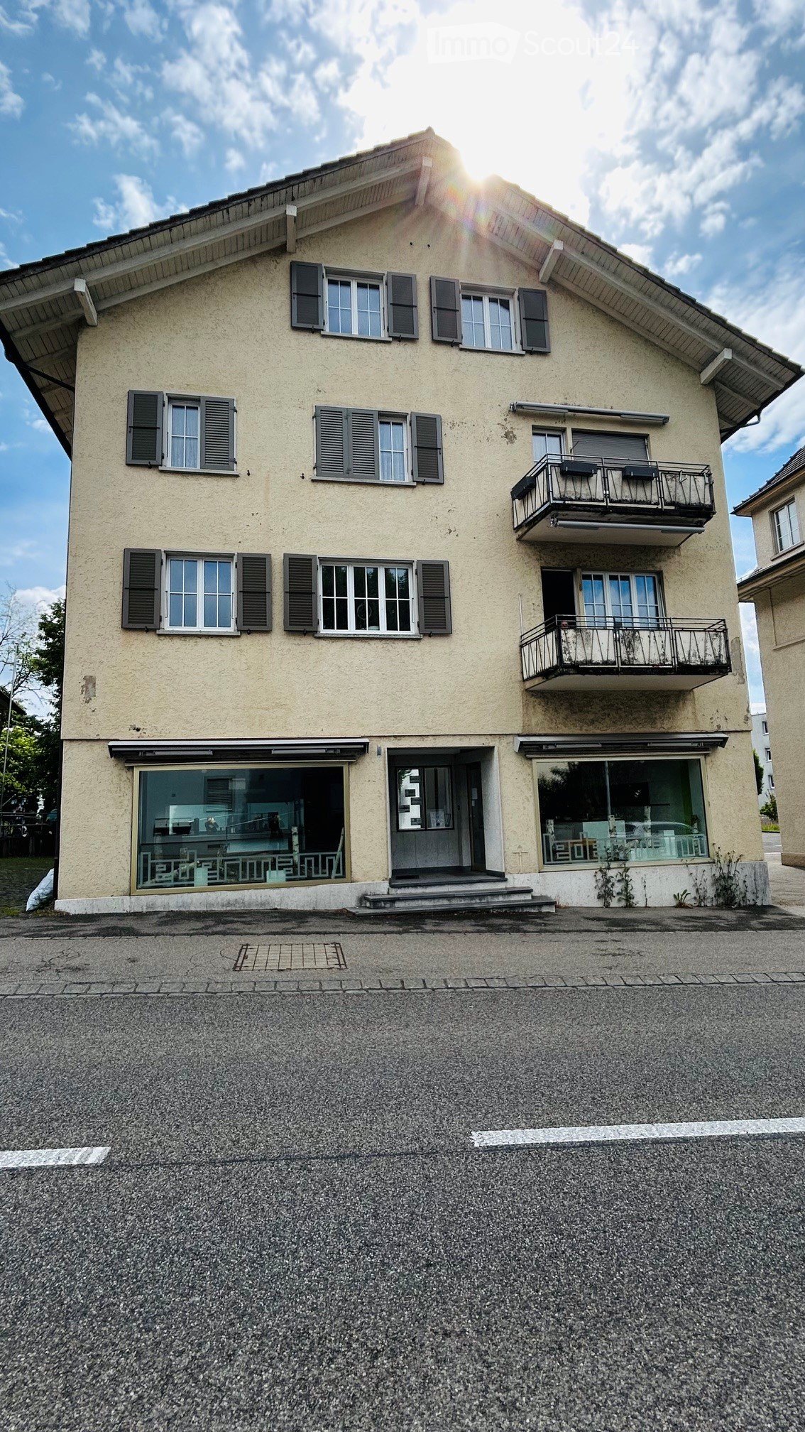 Two-story house with beige exterior, multiple windows with shutters, balconies on the upper floor, and a small entrance area with steps.