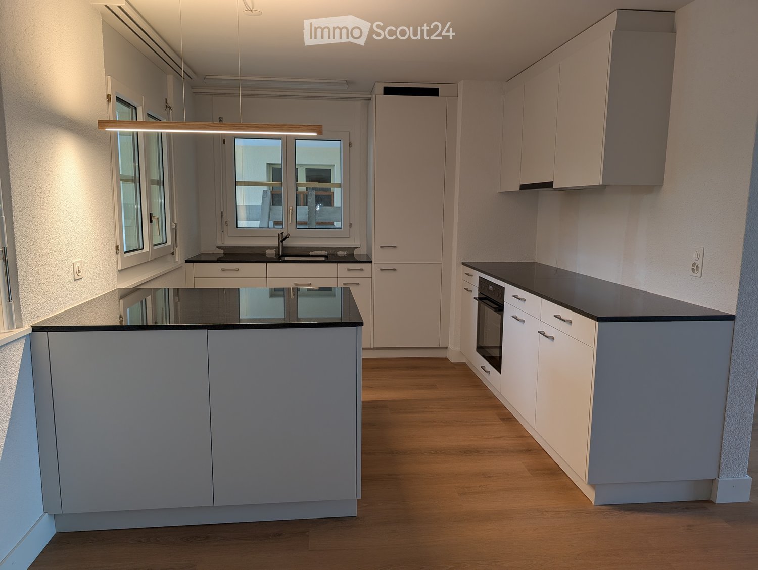Modern kitchen with light grey cabinets, black countertops, a central island, and a sink with a faucet. The space features wooden flooring and large windows.