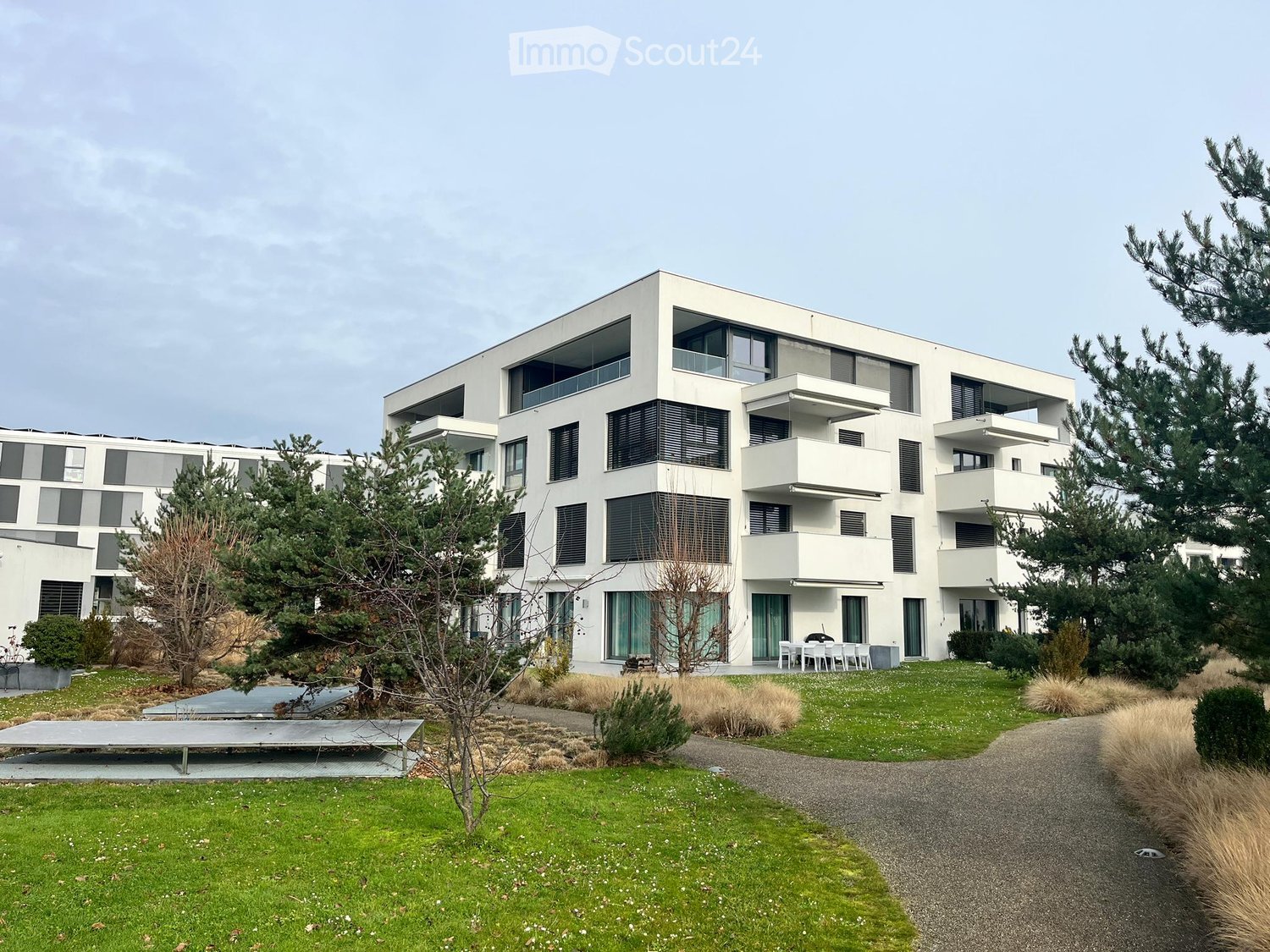 Modern, multi-story residential building with white facade, large windows, and balconies. The surrounding area includes a well-maintained lawn, gravel pathways, and some trees and shrubs.