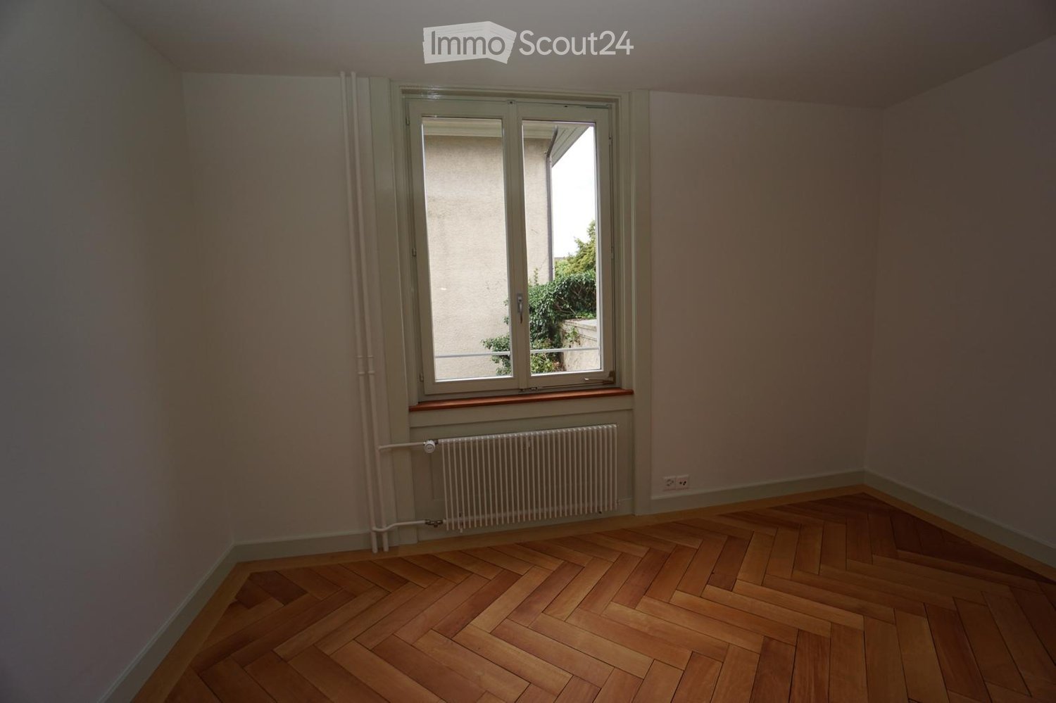 Empty room with wooden herringbone flooring, light-colored walls, a window with a radiator below, and a power outlet visible on the wall.