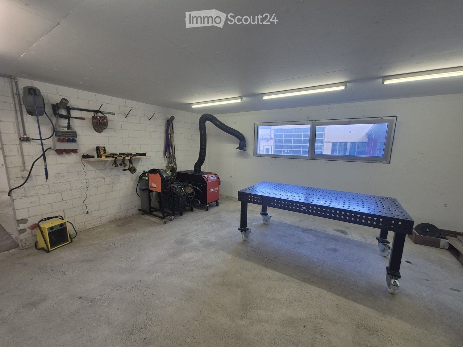 Workshop with a metal workbench, welding machine, power tools, shelves, and a helmet on the wall.