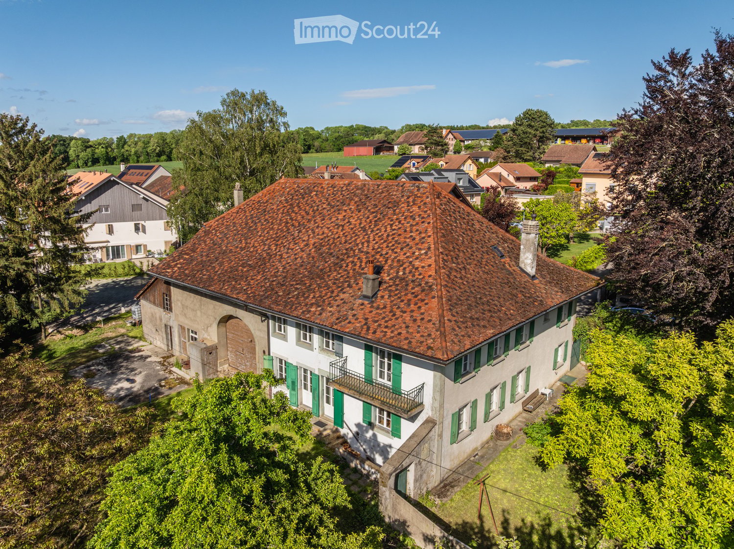 A two-story house with a red-tiled roof, white walls, green shutters, and a small balcony. The house is surrounded by greenery and trees, with neighboring houses visible in the background.