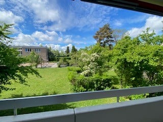 Balcony overlooking a green lawn and trees, with a building visible in the distance under a partly cloudy sky.