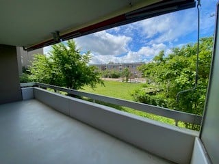 Balcony with white railing, large window offering a view of a green park and buildings.