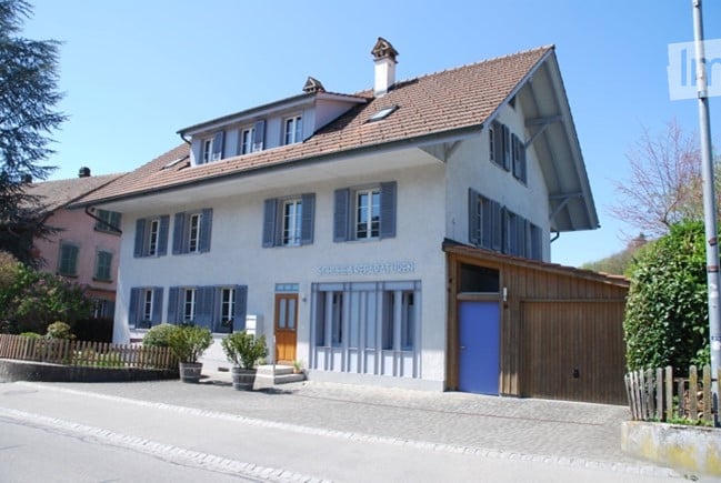 Two-story house with white facade, blue shutters, and a garage. The house has multiple windows and a small garden area in front.