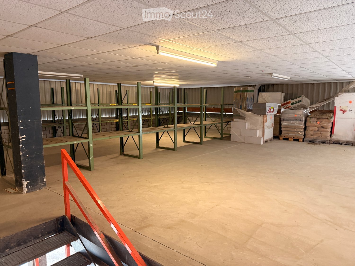 Empty warehouse space with metal shelving units, concrete floor, and exposed ceiling lights. No furniture or equipment present.