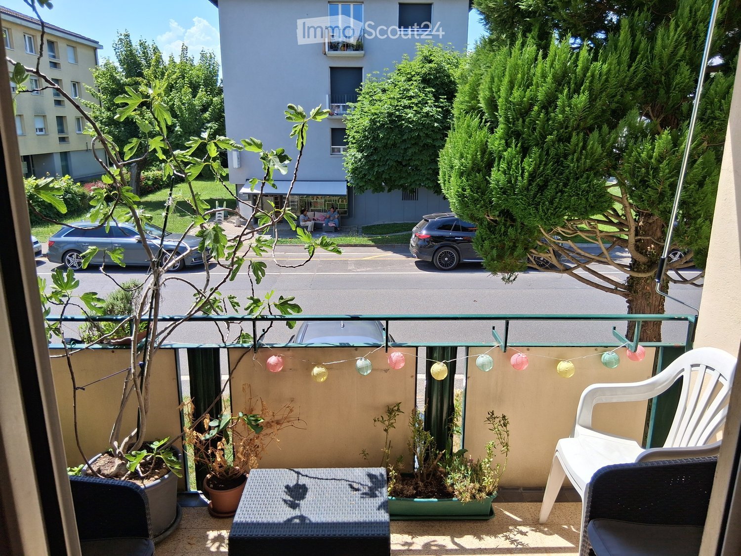 Balcony with a white plastic chair, small table, and several potted plants, with string lights hanging above.