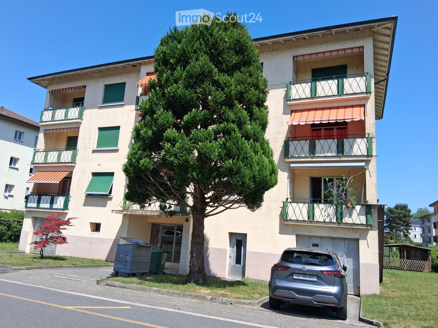 Four-story residential building with balconies, green shutters, and a large tree in front. A silver car is parked in front of the building.