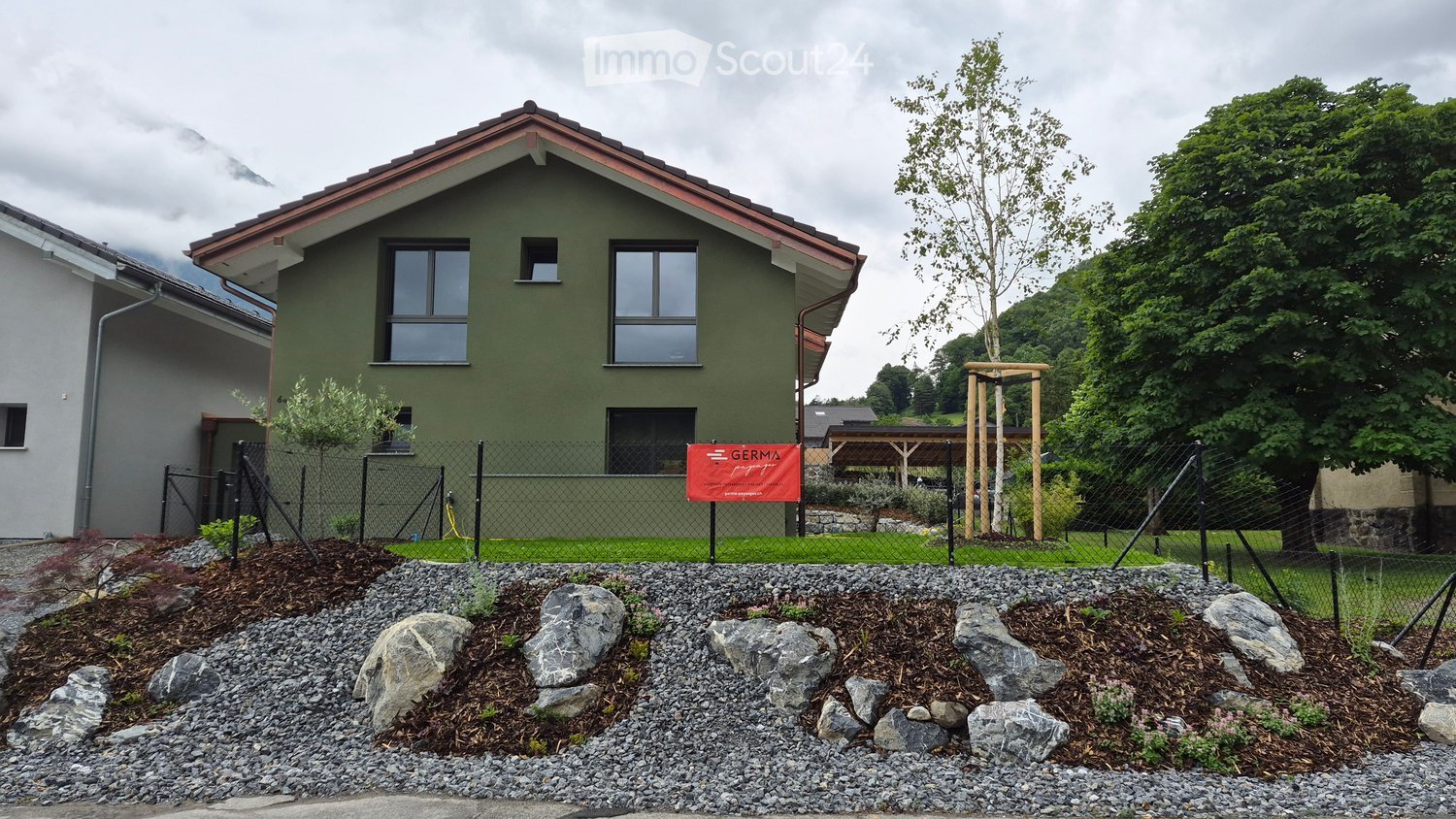 Modern two-story house with green exterior and red roof, surrounded by a landscaped garden with rocks, shrubs, and a fence.