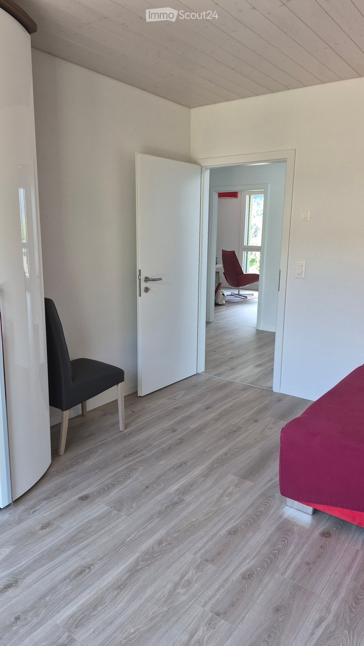 Modern bedroom with light wooden flooring, white walls, and a red bedspread. A black chair is near the door, and an open doorway leads to another room with a red chair and a window.