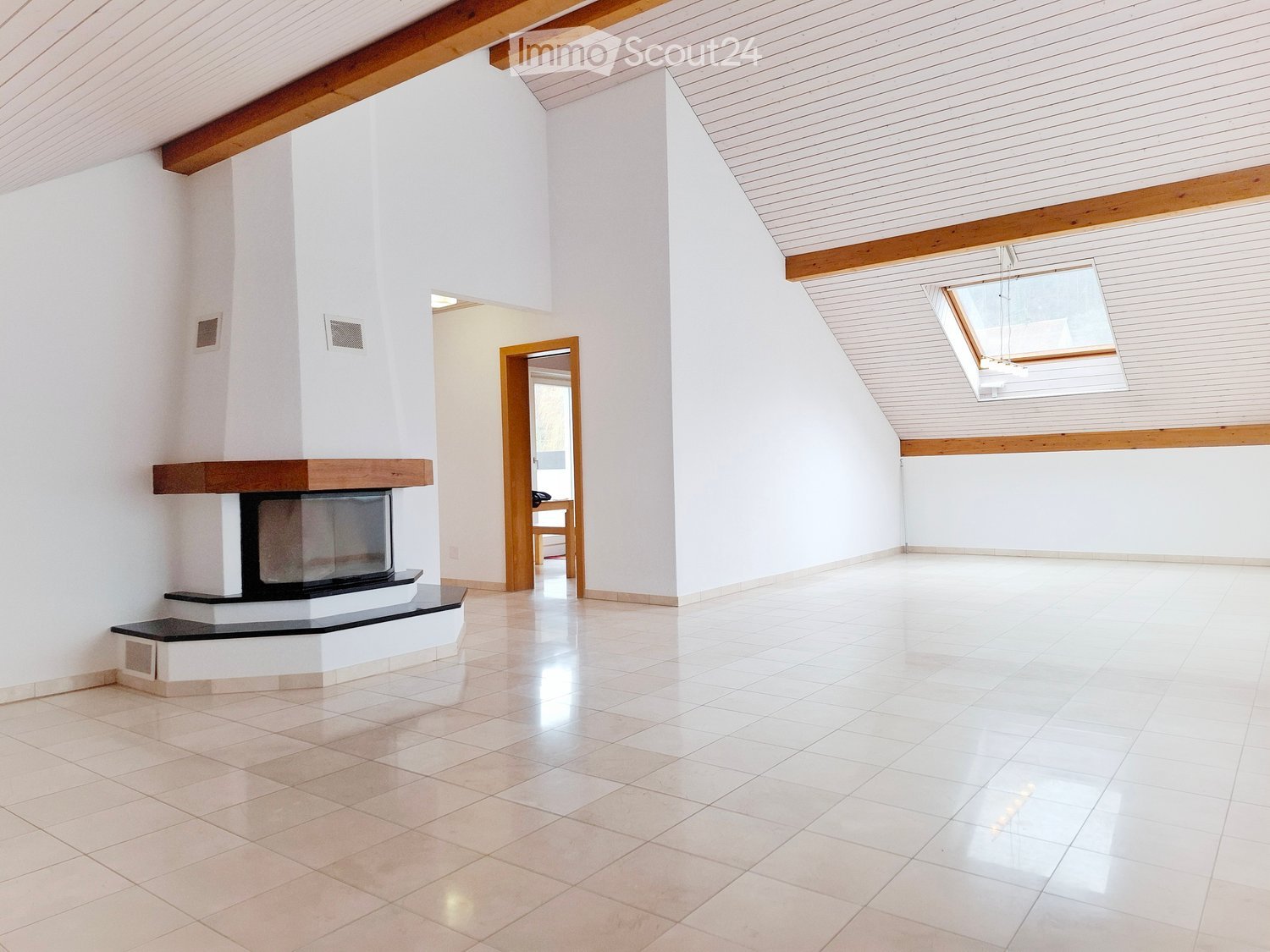 Empty room with white walls, tiled floor, and a built-in fireplace. Wooden beams on the ceiling and a skylight provide natural light.