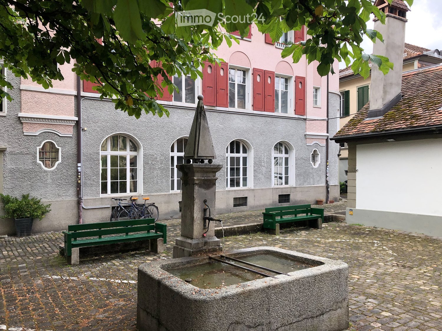 Courtyard with a stone fountain, two green benches, and bicycles leaning against a building.