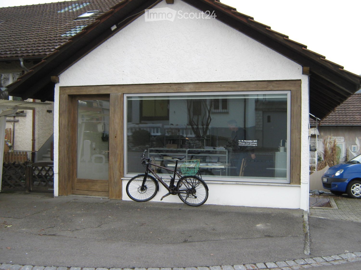 White building with a tiled roof, large window, and wooden door. A bicycle is parked in front, and a blue car is visible to the right.