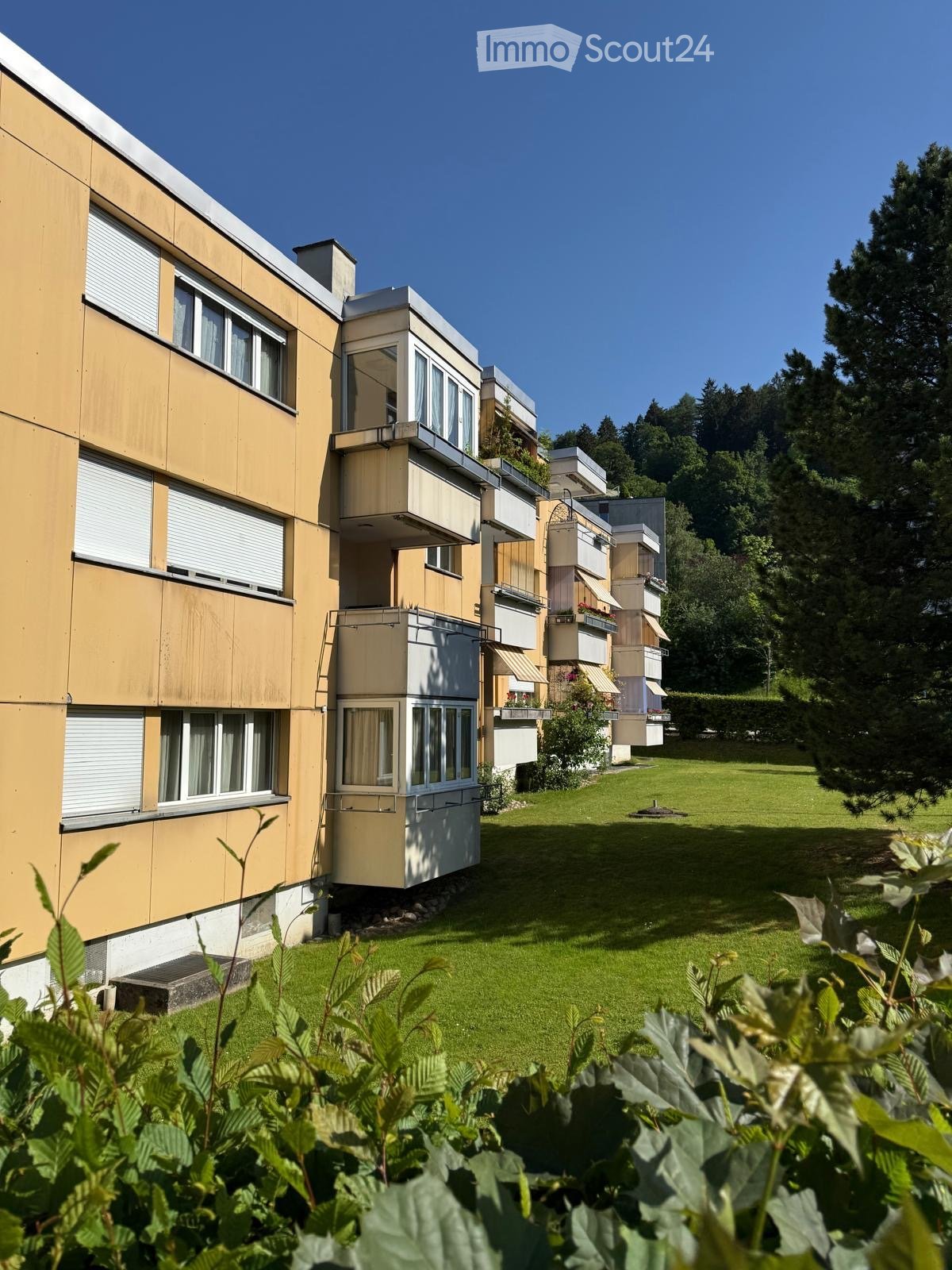 Modern multi-story residential building with balconies and large windows, surrounded by a well-maintained lawn and trees.