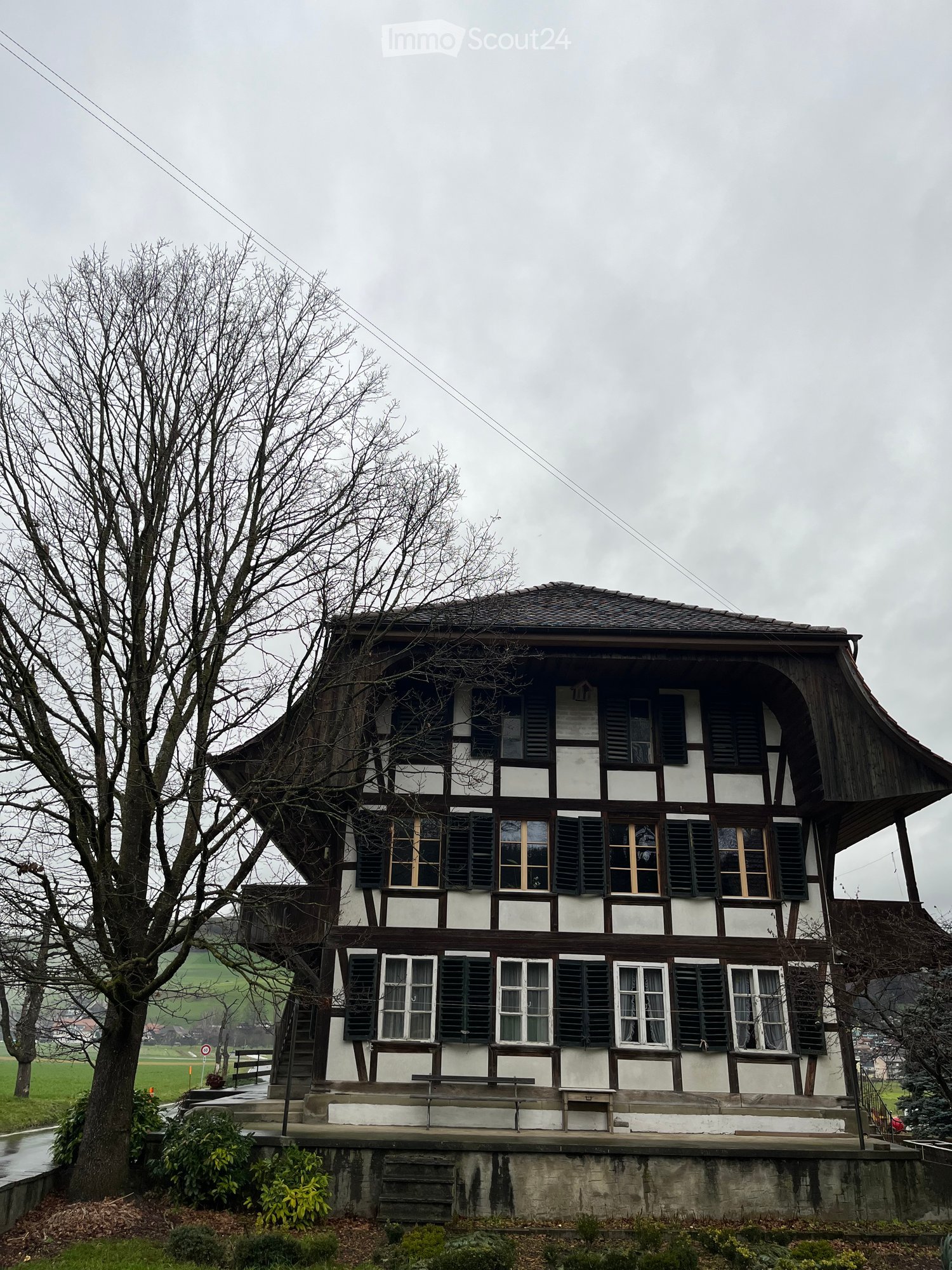 Two-story traditional house with white and dark wooden facade, multiple windows with shutters, and a raised ground floor with a small porch and benches.
