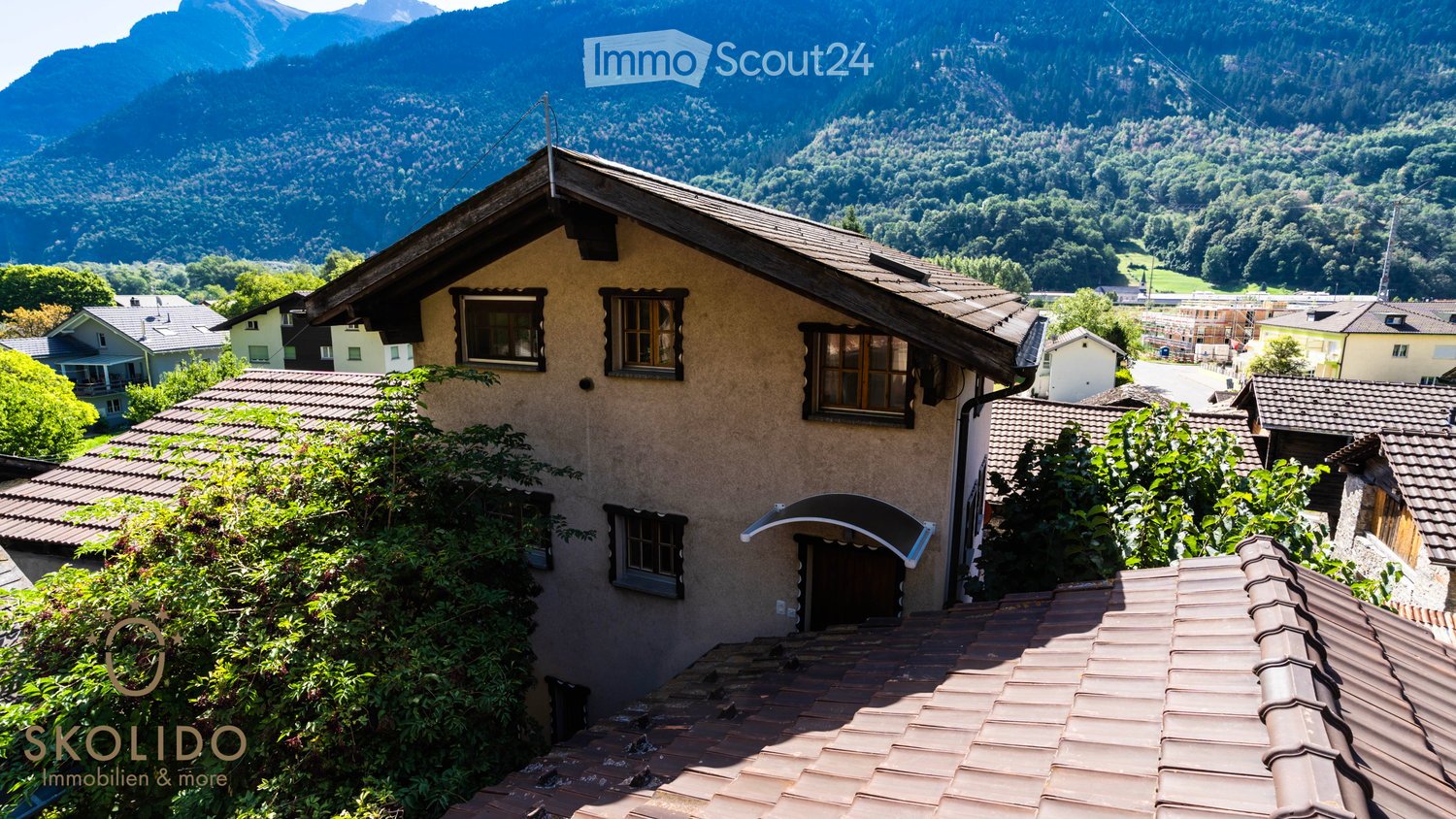 Building. A two-story house with a tiled roof and multiple windows, surrounded by greenery and situated in a scenic mountainous area.
