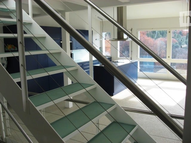 Corridor / Hallway. Modern interior view of a hallway with a staircase leading upwards, featuring glass railings and a sleek design. The area includes a glimpse of a kitchen with blue cabinets and a large window letting in natural light.