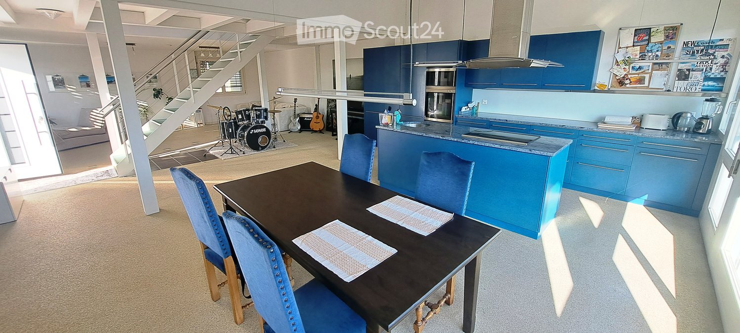 Open-plan living and dining area with a modern kitchen, dining table with chairs, drum set, guitars, and a staircase leading to an upper level.