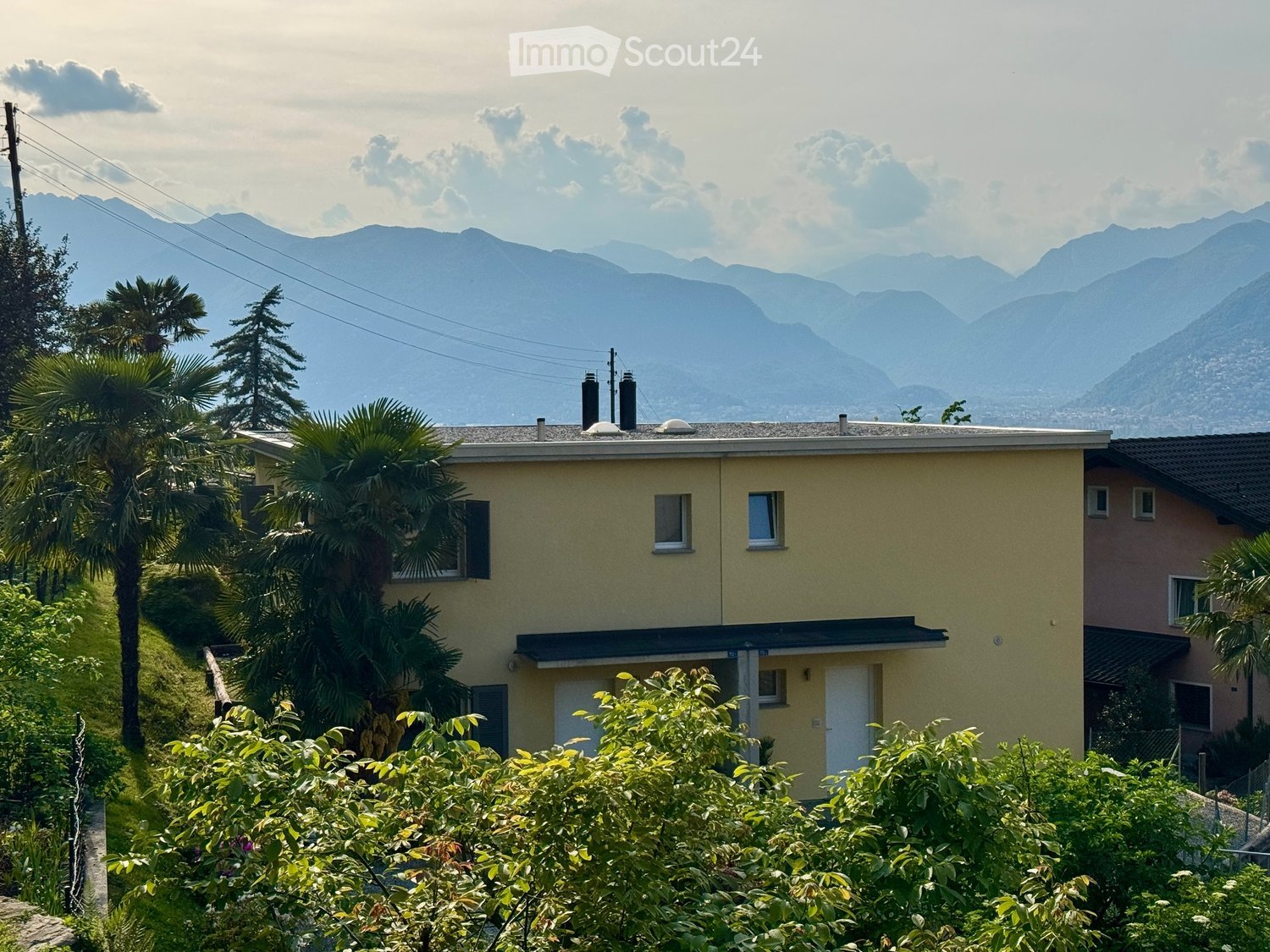 A two-story yellow house with white-framed windows and a gray roof, surrounded by lush greenery and palm trees, with a scenic mountain backdrop.