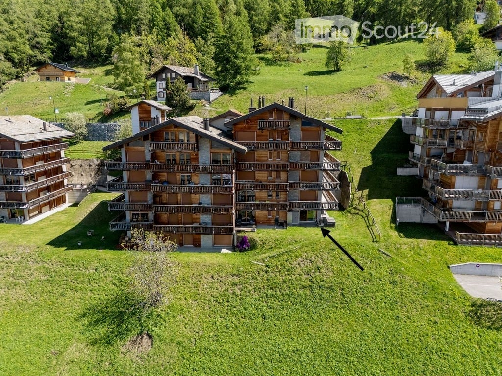 Aerial view of a multi-story wooden apartment building with multiple balconies, surrounded by green hills and other houses.