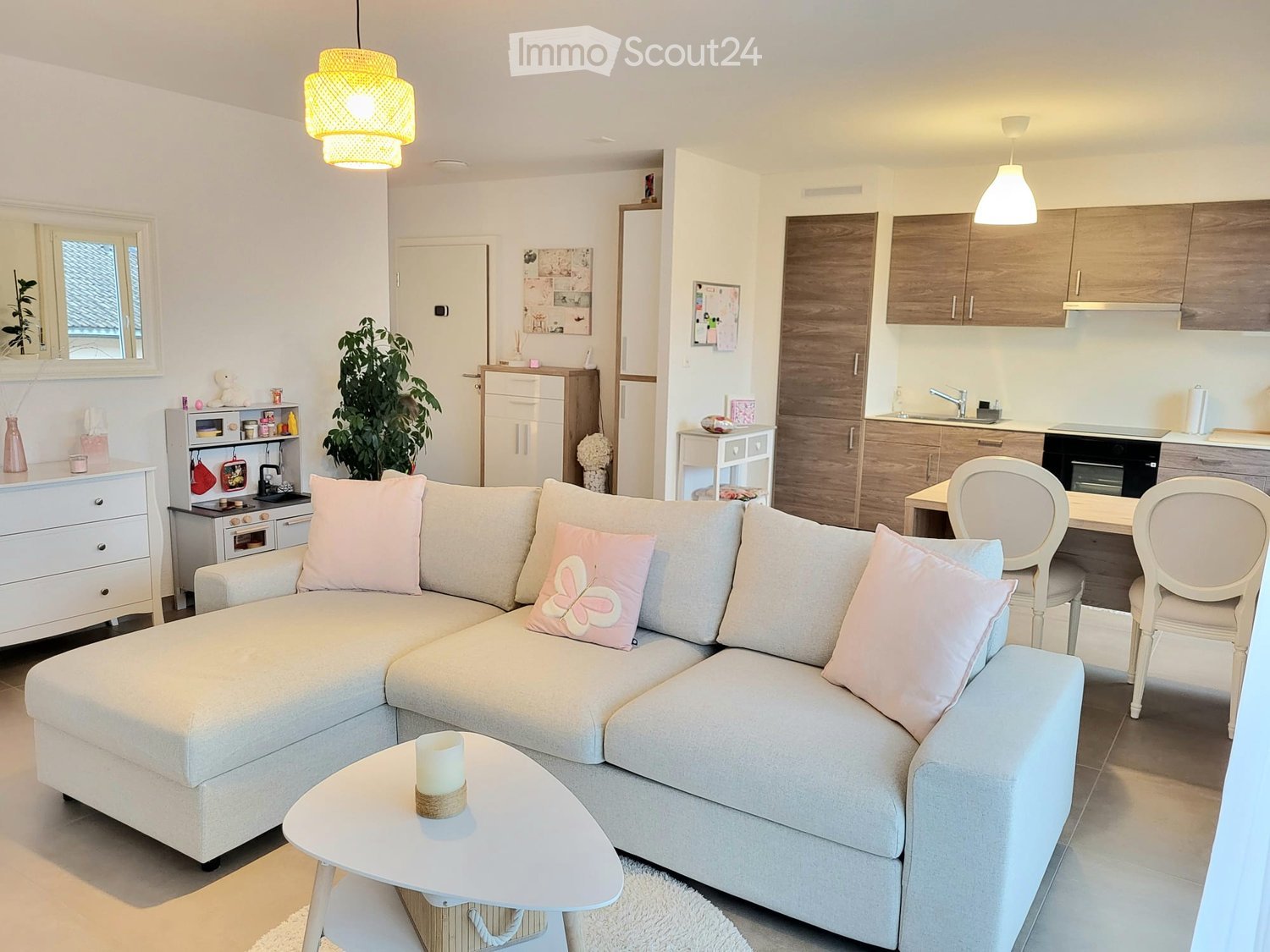 A modern, open-concept living and dining area with a grey sectional sofa, pink cushions, a small coffee table, and a white dining table with two chairs. The kitchen features wooden cabinets, a sink, and a small stove.
