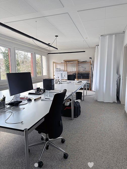 Modern office space with large windows, white walls, and a grey carpeted floor. A white desk holds a computer monitor, keyboard, mouse, and office supplies. An office chair is positioned in front of the desk. A black curtain is on the right side, and shel