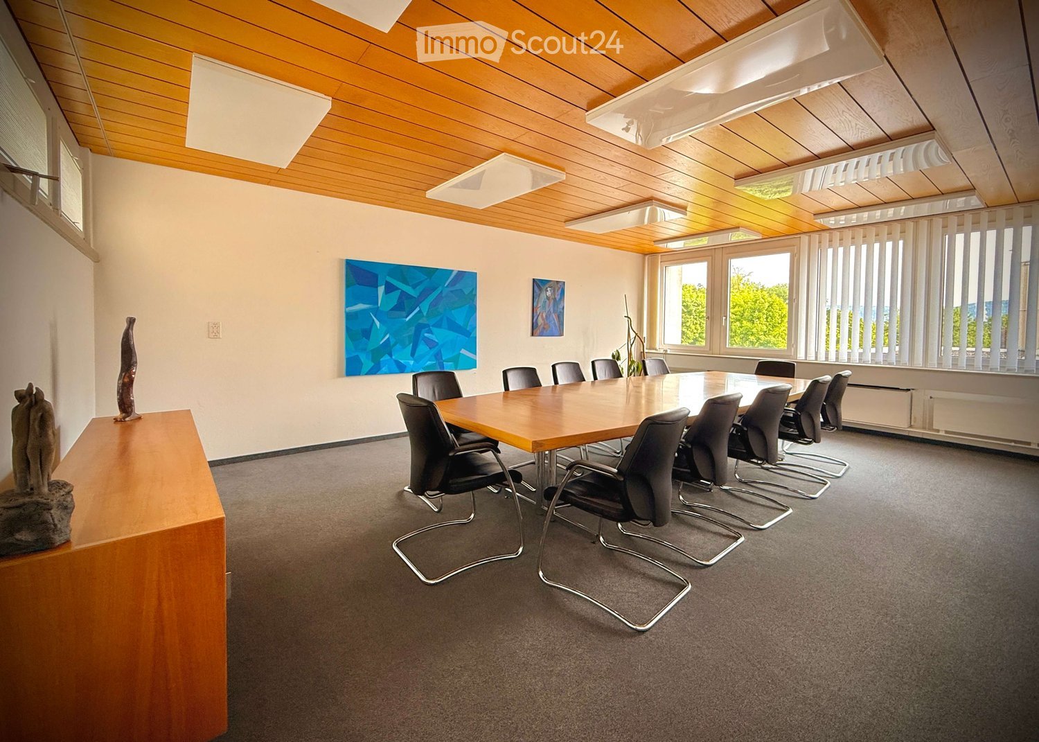 Conference room with a long wooden table surrounded by black chairs, a sculpture on a side table, and artwork on the walls. Large windows provide natural light.