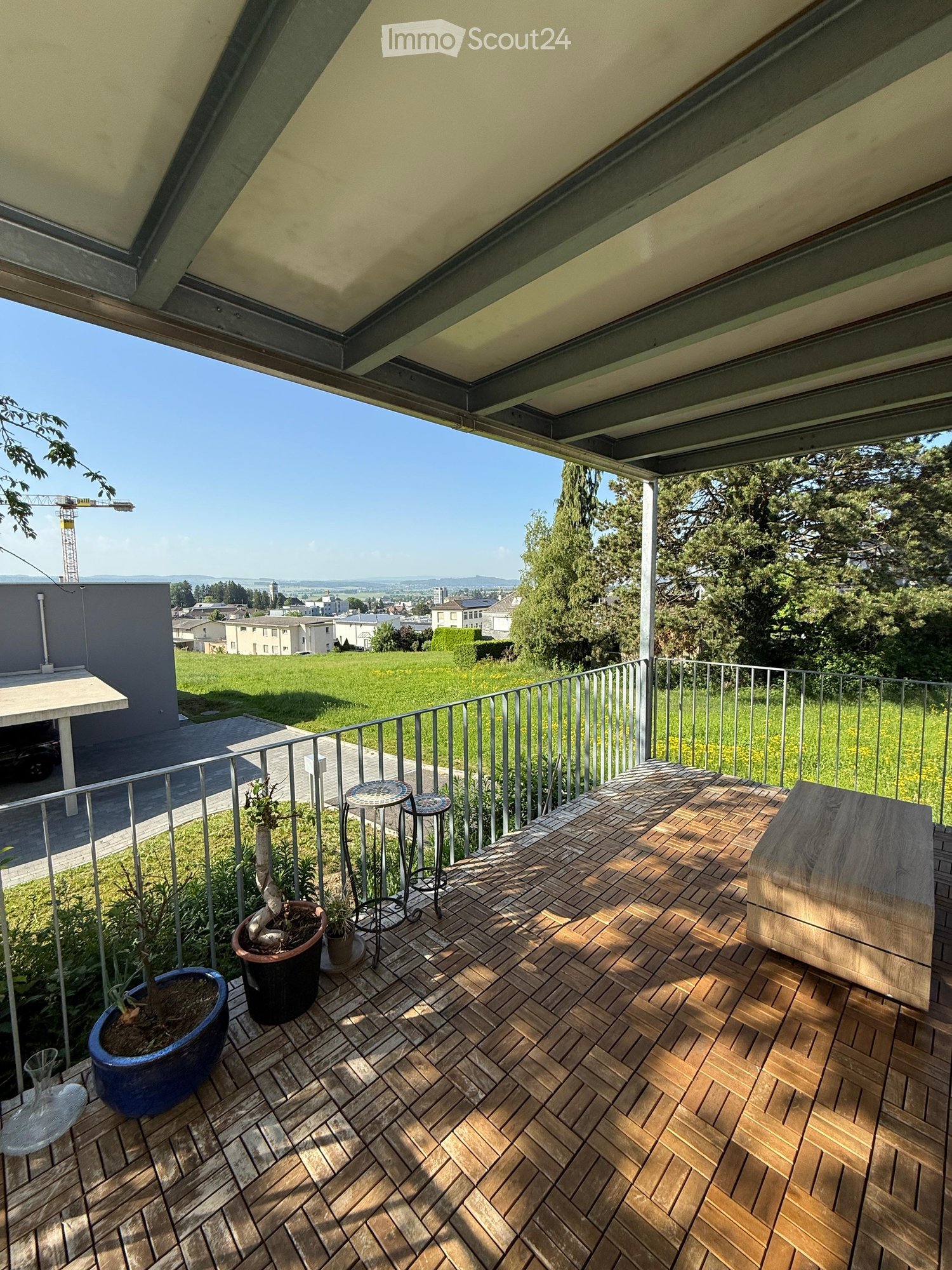 Wooden decked balcony with a small table, stools, and potted plants, overlooking a green landscape and town.
