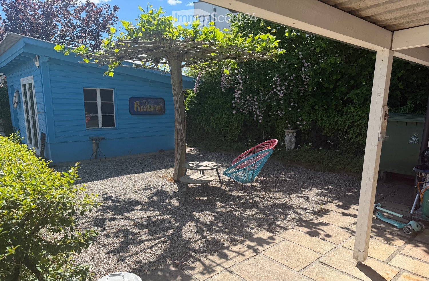 A small outdoor seating area with a table, chairs, and a stool, surrounded by greenery and a blue building.