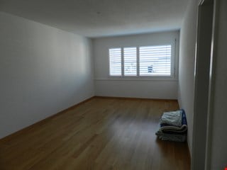 Empty room with wooden flooring, white walls, and a single window with blinds. A folded blanket is placed in the corner.