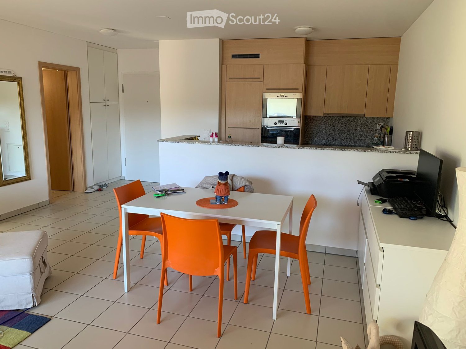 A modern, well-lit room with a white dining table and orange chairs, a built-in kitchen area with cabinets and an oven, a white sofa, and a mirror on the wall.