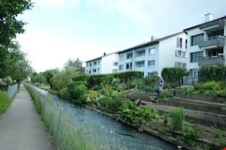 A narrow garden path runs alongside a small water channel, bordered by low shrubs and trees. A person is walking along the path, and modern apartment buildings are visible in the background.