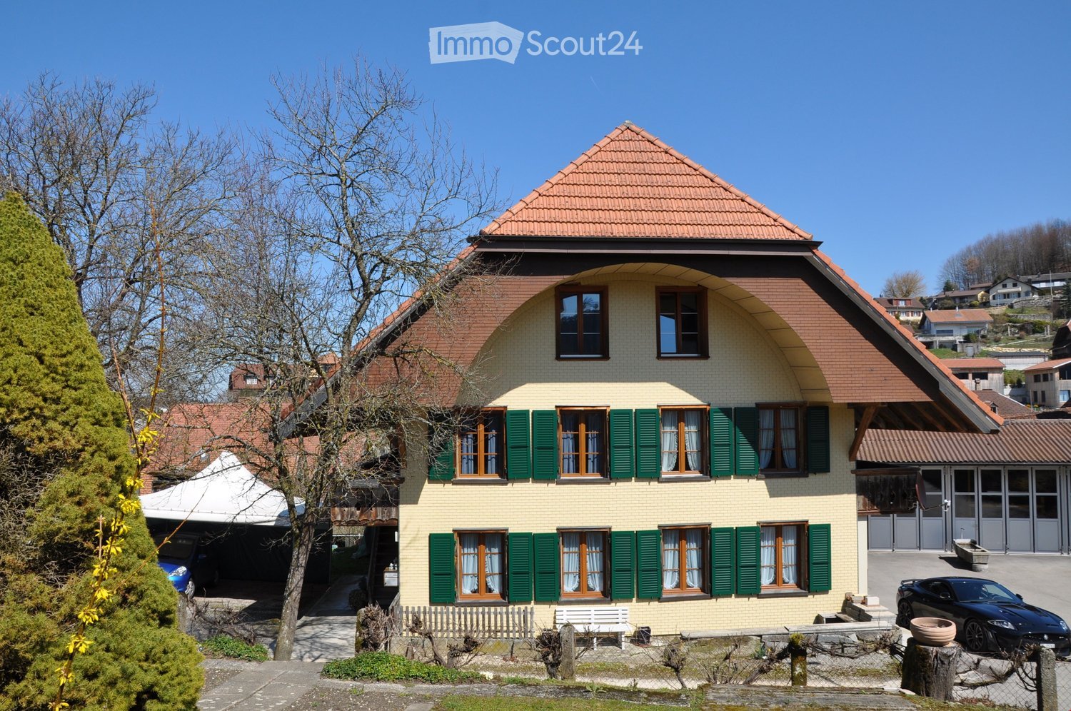 Two-story house with a red-tiled roof, green shutters, and multiple windows. The house features a garage and is surrounded by trees and a small garden area.