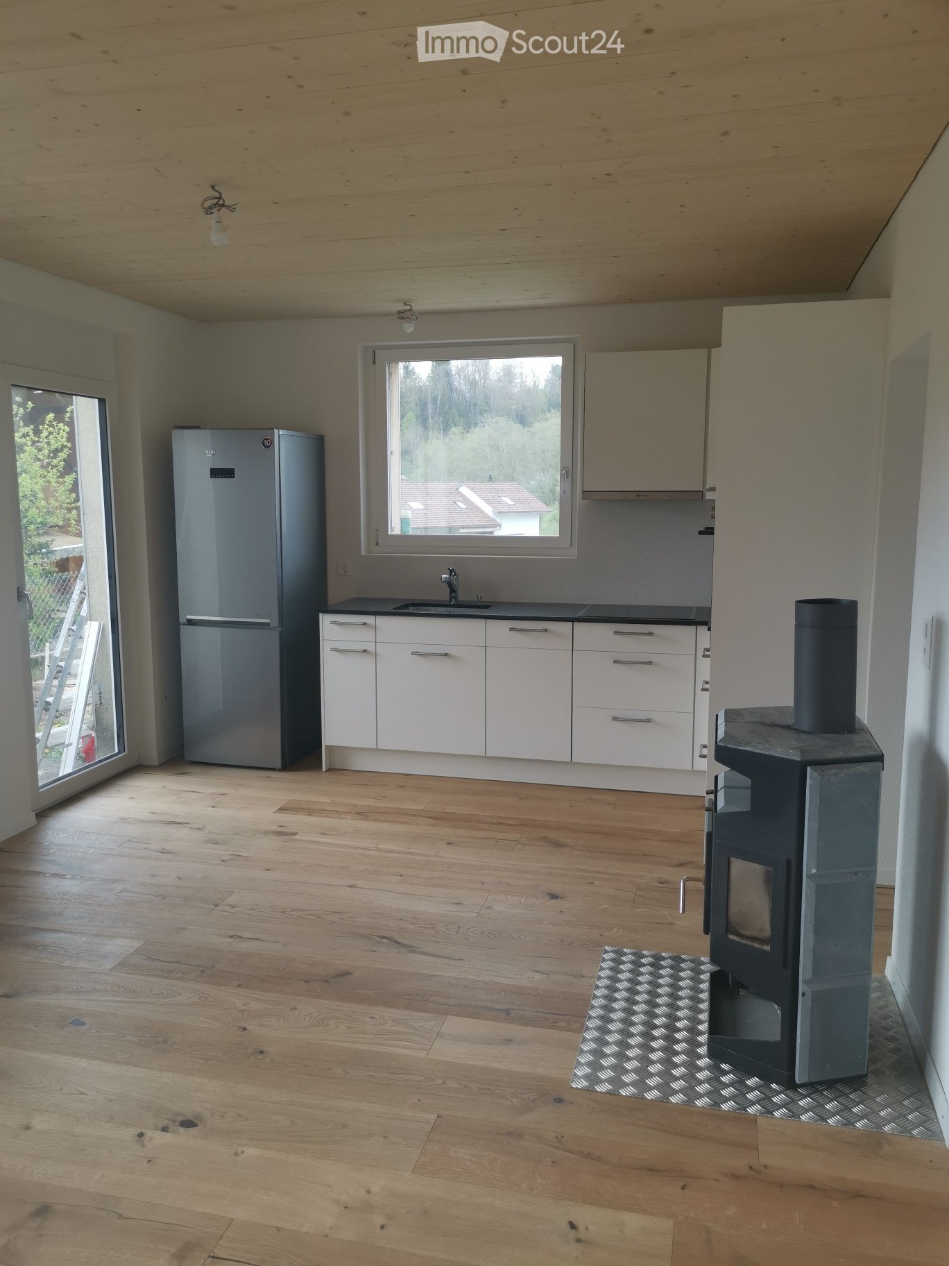Modern kitchen with light wooden flooring, white cabinets, stainless steel refrigerator, and a wood stove with a patterned rug in front.